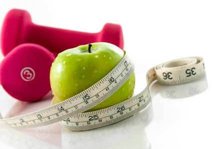 Fresh appetizing apple and brightly colored dumbbells tied with a measuring tape. Slight reflection, white background, focus on the apple