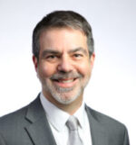 Head and shoulders of man wearing white shirt light gray tie, medium gray suit jacket, gray facial hair and short gray and black hair smiling