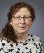 head and shoulders of owman with dark shoulder length curly hair, dark framed eyeglasses and geometric patterned shirt in black, blue white and yellow/green