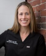 Woman leaning against brick wall wearing dark scrubs smiling