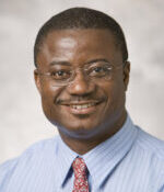 head and shoulders of man with dark short hair, wearing frameless eyeglasses, blue shirt, red tie