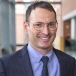 Man with brown hair, eyeglasses, blue suit jacket, shirt and tie, smiling