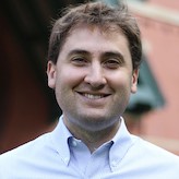 head and shoulders of man with brown hair, white button down shirt, smiling broadly against background of outside of brick building