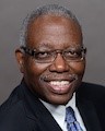 Head and shoulders of man with short gray hair, eyeglasses, dark suit jacket, light shirt, blue necktie, smiling broadly