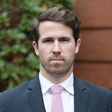 head and shoulders of man with brown hair, white shirt, pink tie, blue/gray suit jacket, neutral expression, against background of green shrub and brick building