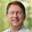 Man with brown hair wearing frameless eyeglasses wearing light colored open neck shirt smiling