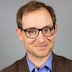 man with brown hair, dark frame eyeglasses, blue shirt, brown jacket