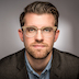 Man with brown hair, clear frame glasses, blue shirt, bown jacket