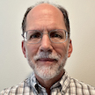 head and shoulders of man with gray hair and facial hair, light colored plaid shirt, frameless eyeglasses