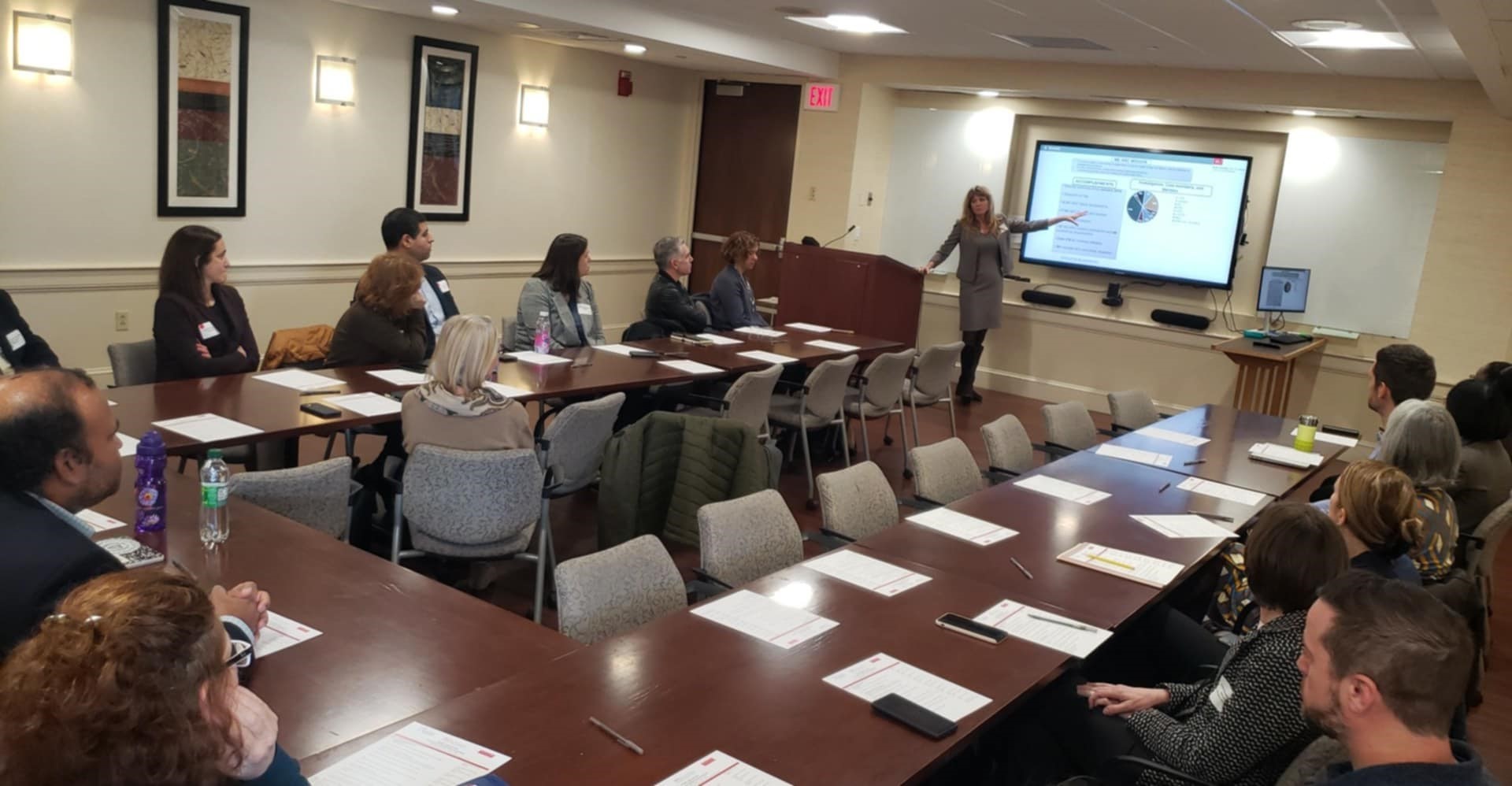 People seated around large U shaped table wooden table watching a woman present slides on a digital screen