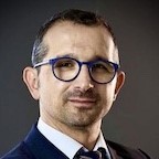 Headshot of Alexandros Tsouknidas dark short hair dark round eyeglasses, white shirt, dark suit jacket
