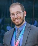 man wearing gray shit and blue shirt, brown hair, facial hair, eyeglasses, smiling